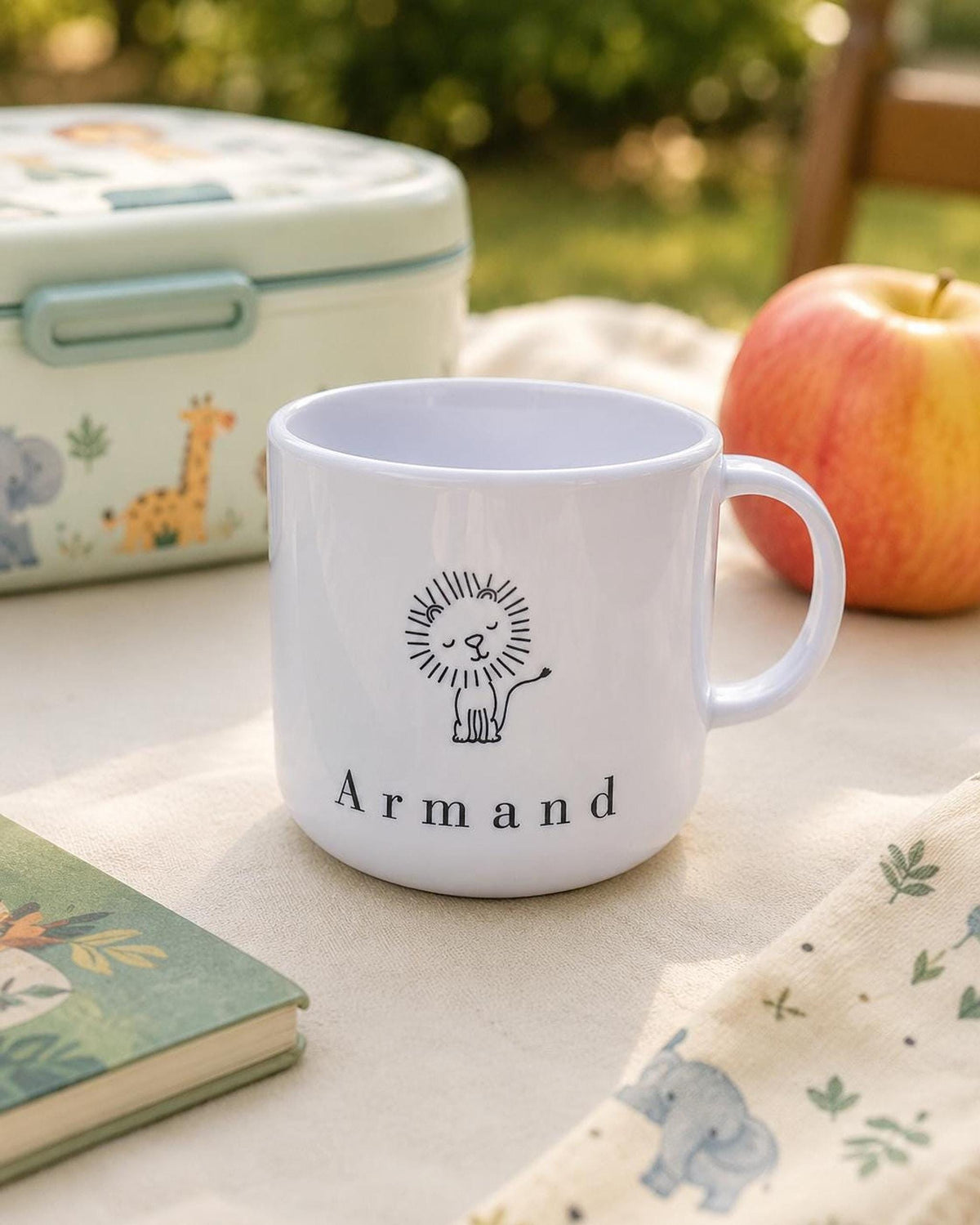 The personalized white lion mug is set outdoors on a picnic table next to a red apple, an illustrated lunchbox, and a book.