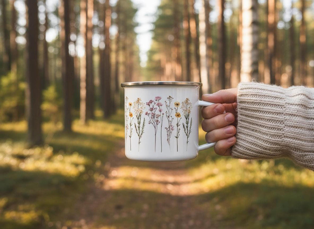 A person wearing a cream knit sweater holds the wildflower enamel mug outdoors in a sunlit pine forest.