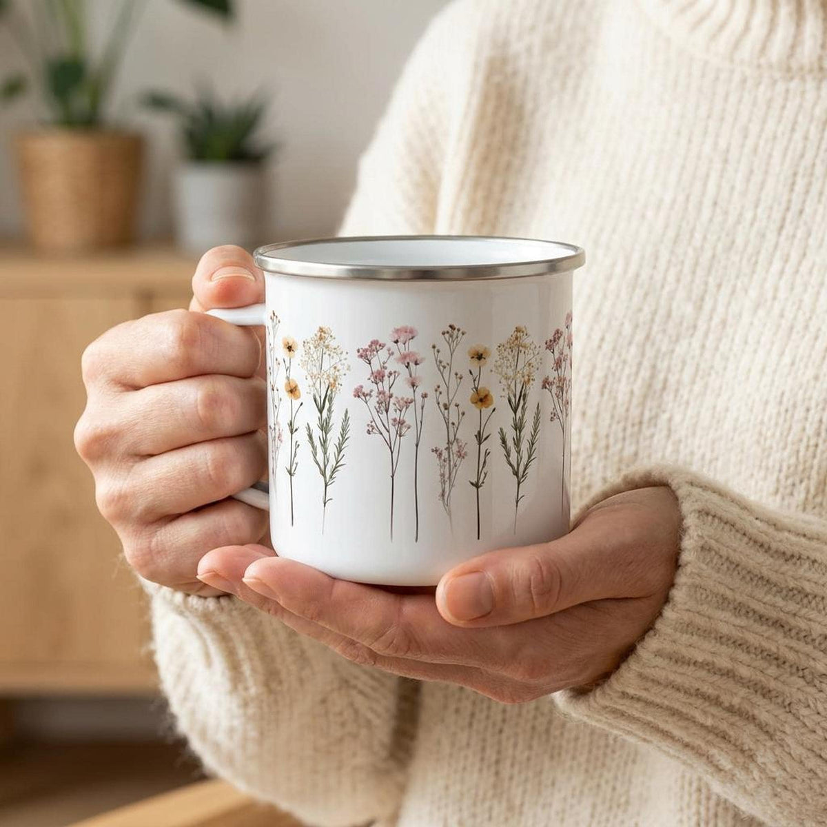 A person wearing a cream sweater holds the enamel mug with both hands in a bright indoor room.