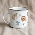 A white enamel mug with a silver rim and handle, printed with colorful safari animals including a leopard, lion, and koala, sitting on a natural beige cloth surface.