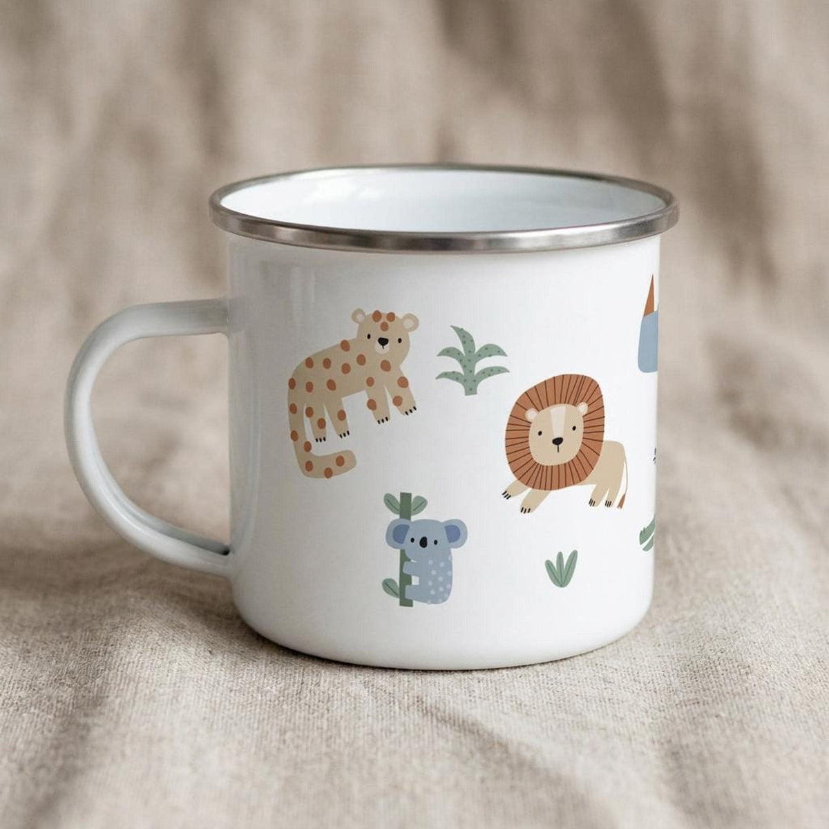 A white enamel mug with a silver rim and handle, printed with colorful safari animals including a leopard, lion, and koala, sitting on a natural beige cloth surface.