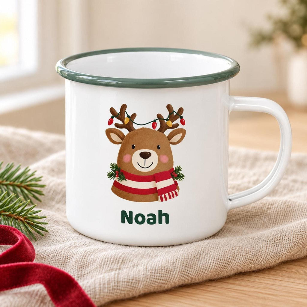 A white enamel mug with a green rim features an illustration of a reindeer with string lights on its antlers, wearing a striped scarf, and the name Noah printed underneath.