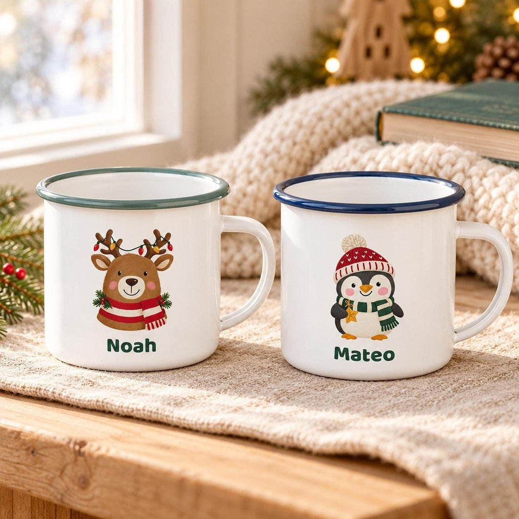 Two white enamel mugs sit side-by-side on a neutral cloth, one with a green rim showing a reindeer and the name Noah, and one with a blue rim showing a penguin and the name Mateo.