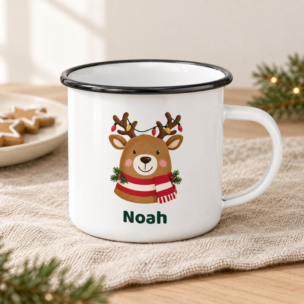 A white enamel mug with a black rim displays a reindeer graphic with the name Noah, set on a beige cloth next to a plate of star-shaped cookies.