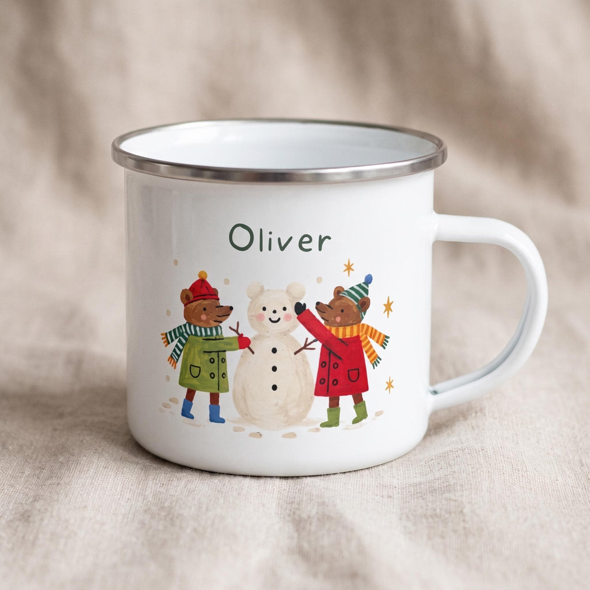 A white enamel mug with a silver rim displaying the name Oliver above an illustration of two bears building a snowman in winter clothing.