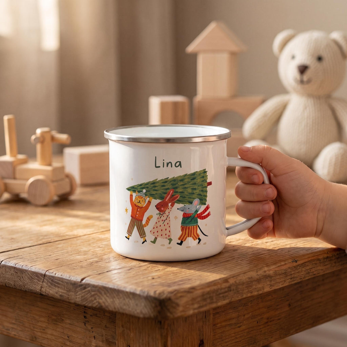 A child's hand holds a personalized white enamel mug with a silver rim and an illustration of animal characters carrying a Christmas tree, sitting on a wooden table with toys.