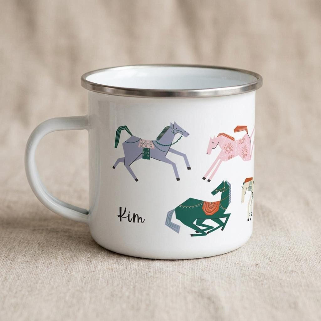 A white enamel mug with a silver rim displays colourful stylized horse illustrations and the name Kim on a beige fabric background.
