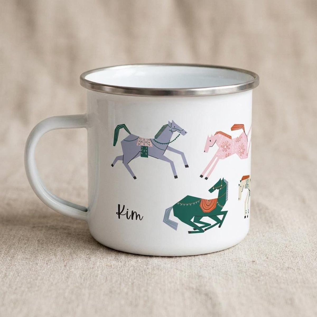 A white enamel mug with a silver rim displays colourful stylized horse illustrations and the name Kim on a beige fabric background.