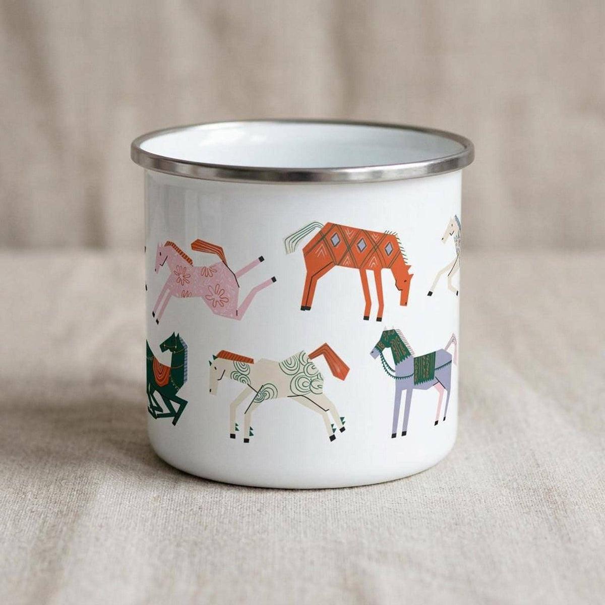 A white enamel mug with a silver rim sits on a textured fabric surface, showing an alternate side with printed folk-art horses.