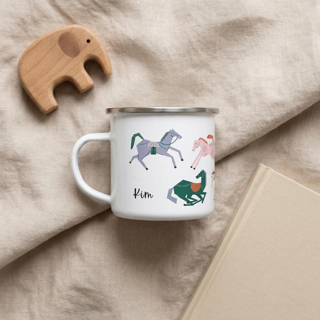 A white enamel mug with horse graphics and the name Kim rests on a beige blanket next to a wooden elephant toy and a closed book.
