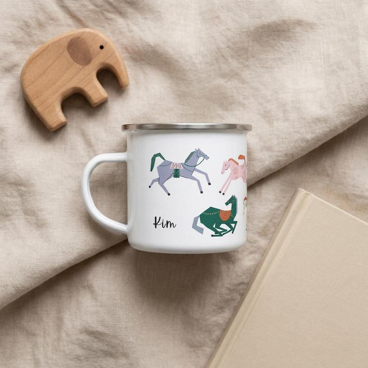 A white enamel mug with horse graphics and the name Kim rests on a beige blanket next to a wooden elephant toy and a closed book.