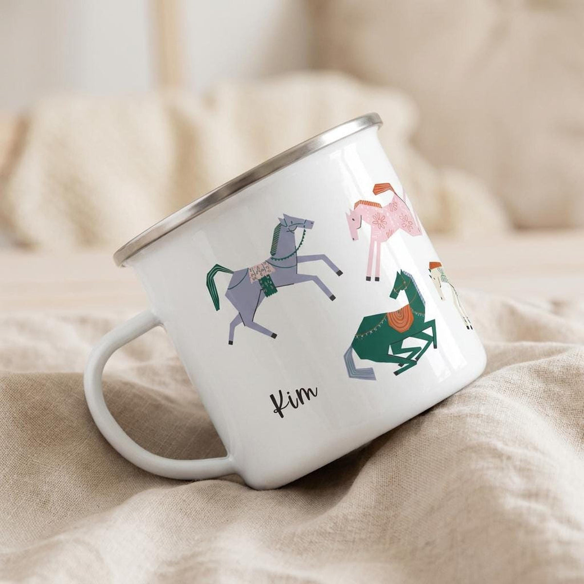 A white enamel mug with cartoon horse designs and the name Kim rests tilted on a light neutral cloth surface.