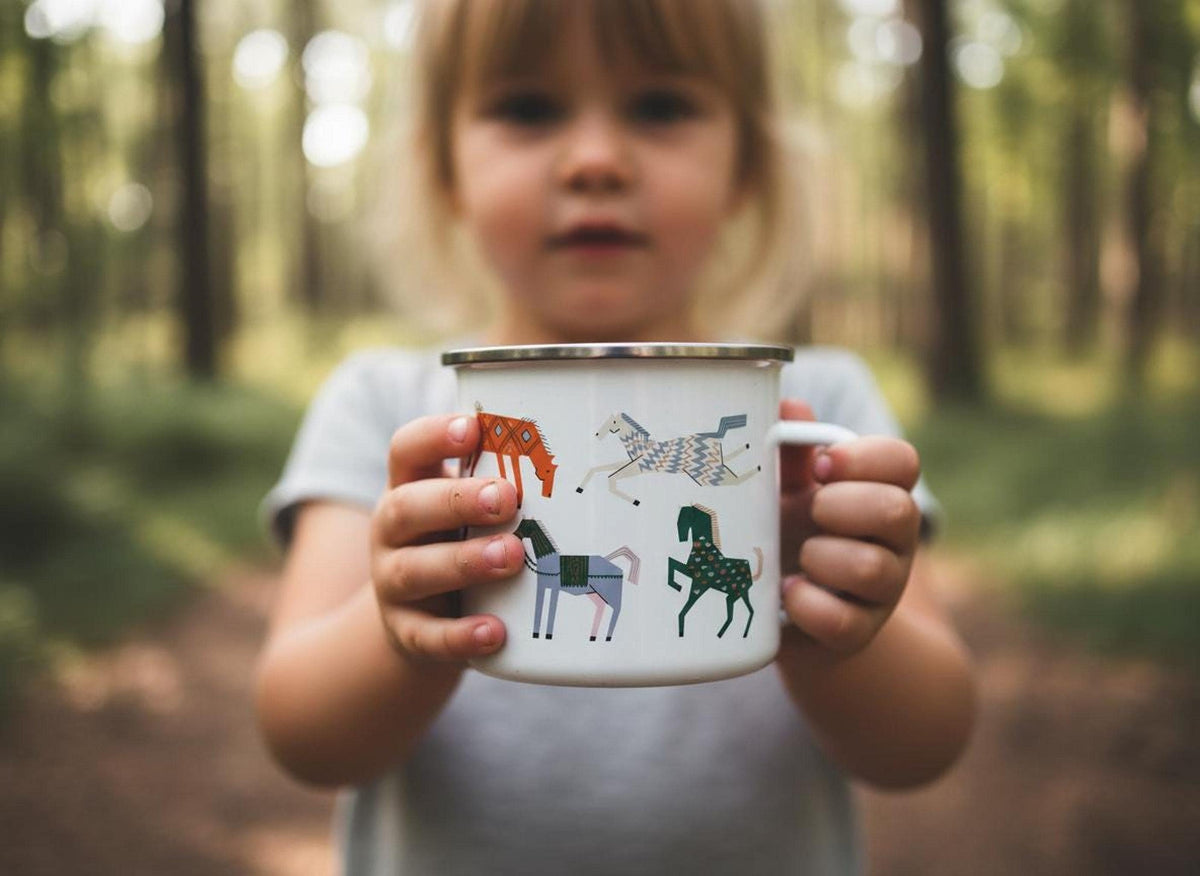 A young child holds out a white enamel mug decorated with colourful horse illustrations in front of a blurred forest background.