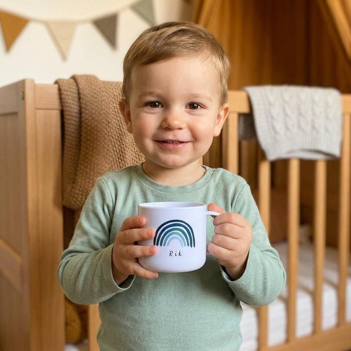 A smiling young child in a green long-sleeve shirt holds a white mug with a blue rainbow graphic and the name Rik.