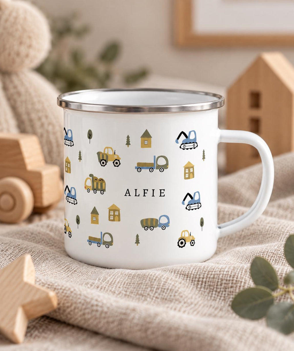 A white enamel mug with a silver rim and a handle, featuring a repeating pattern of small construction vehicles, houses, and trees in muted tones. The name ALFIE is printed in black capital letters in the centre of the mug.