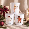 Three white cups featuring animal illustrations and names. The top cup shows a rabbit labelled NORA. The bottom left cup shows a dog playing football labelled HUGO. The bottom right cup shows a bear skiing labelled ELIO.