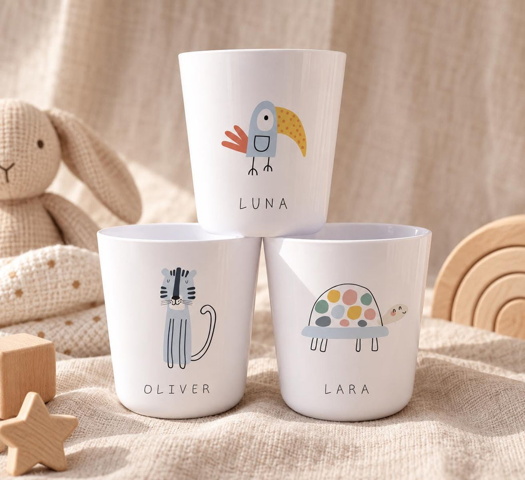 Three white cups featuring colourful animal illustrations and personalised names. The top cup displays a bird and the name LUNA. The bottom left cup shows a tiger and the name OLIVER. The bottom right cup features a turtle and the name LARA.