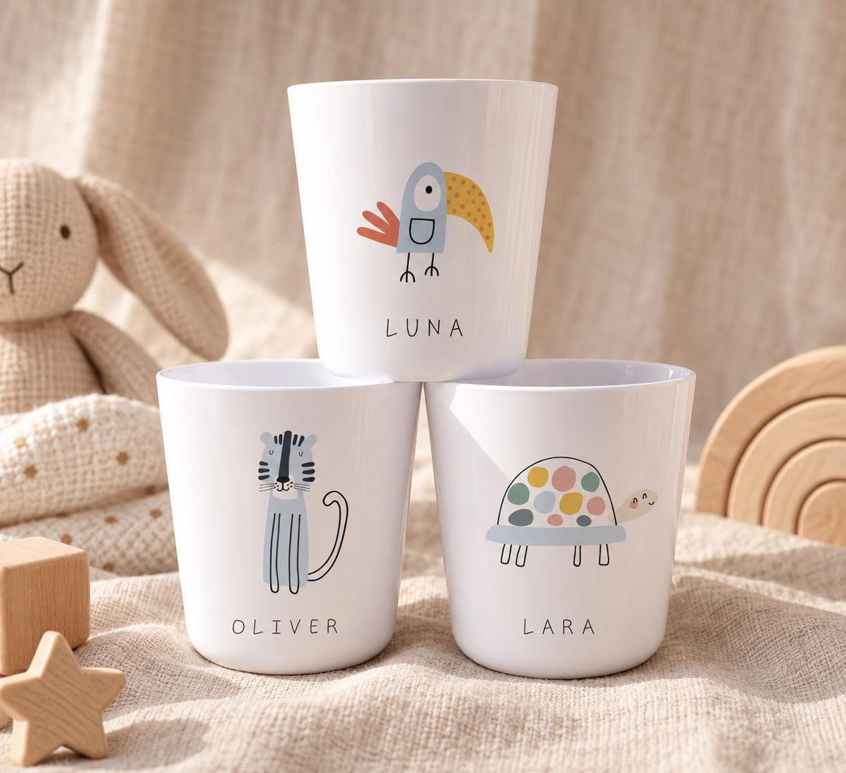 Three white cups featuring colourful animal illustrations and personalised names. The top cup displays a bird and the name LUNA. The bottom left cup shows a tiger and the name OLIVER. The bottom right cup features a turtle and the name LARA.