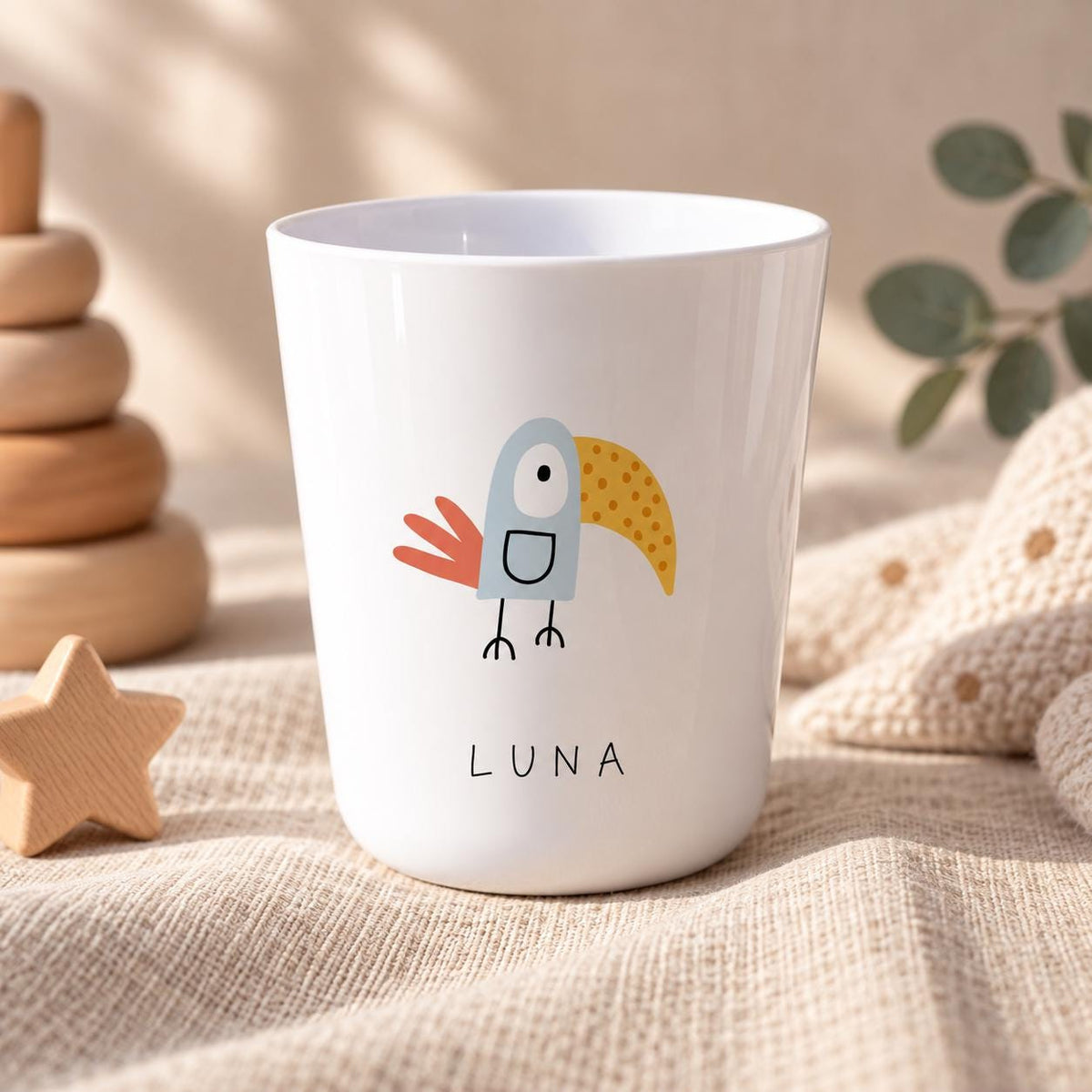 A white drinking cup featuring a stylised illustration of a bird with a light blue body, a yellow dotted beak, and red tail feathers. The name LUNA is printed in black capital letters below the bird graphic on the front of the cup.