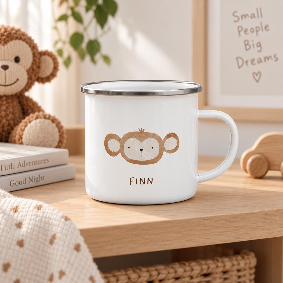 A white enamel mug with a silver rim and a handle, featuring a brown illustrated monkey face and the name FINN printed in brown text below the graphic. The mug is positioned on a wooden surface with a soft, neutral-toned aesthetic.