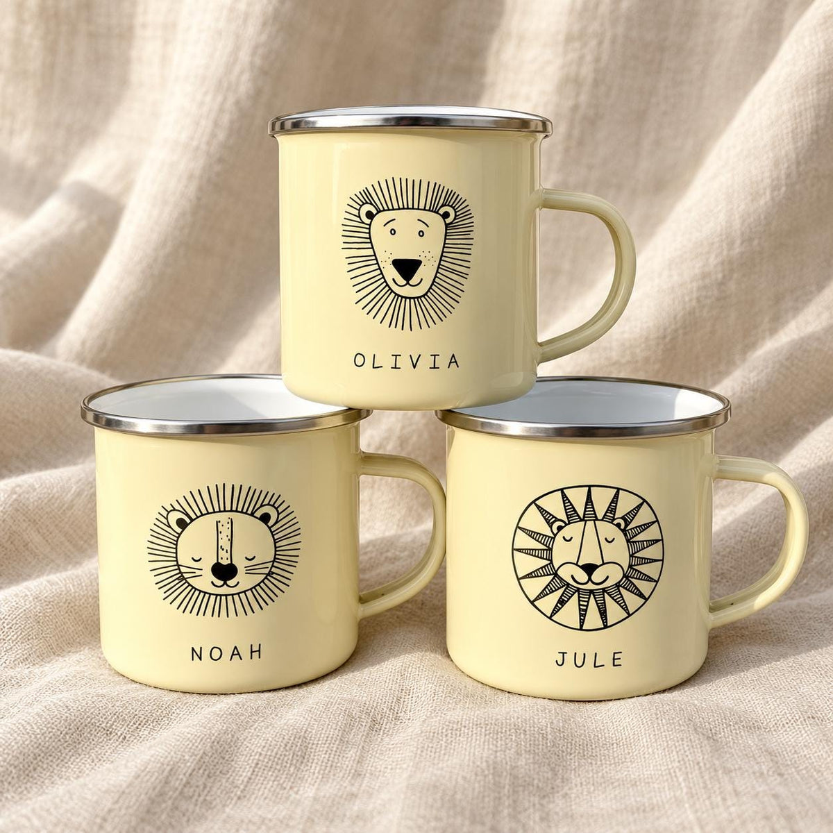 Three pale yellow enamel mugs with silver rims, each featuring a black line-art illustration of a lion face. The mugs are personalised with the names Olivia, Noah, and Jule printed in black capital letters below the lion graphics.