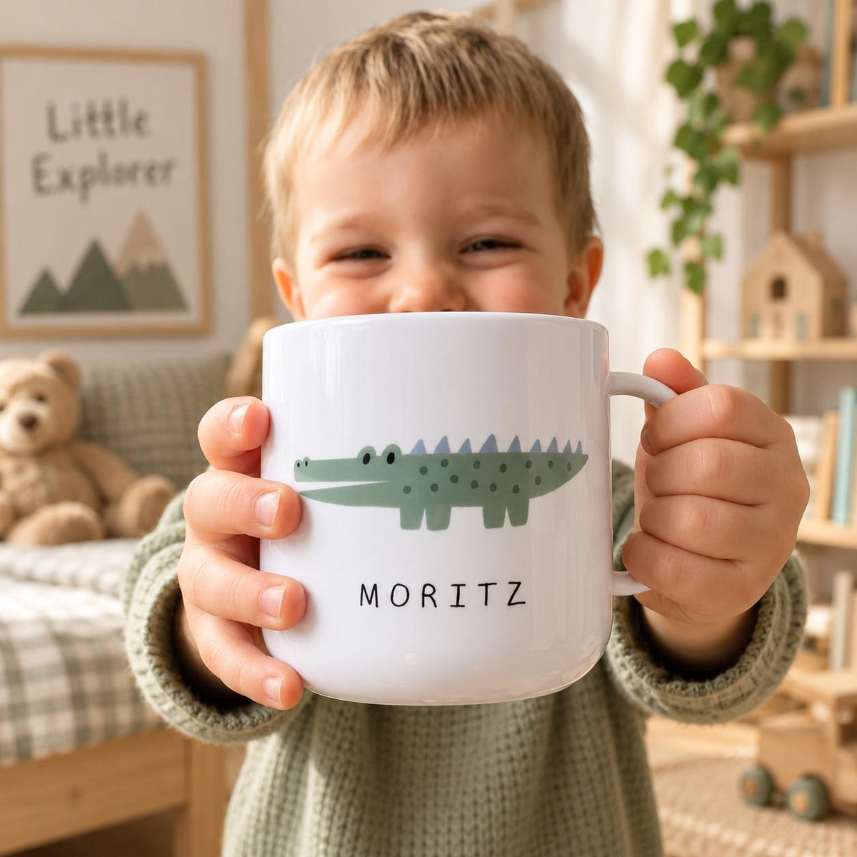 A white kids cup featuring a green crocodile illustration with a light blue spiky back and the name MORITZ printed in black capital letters below the graphic. The cup has a simple handle on the right side and a smooth finish.