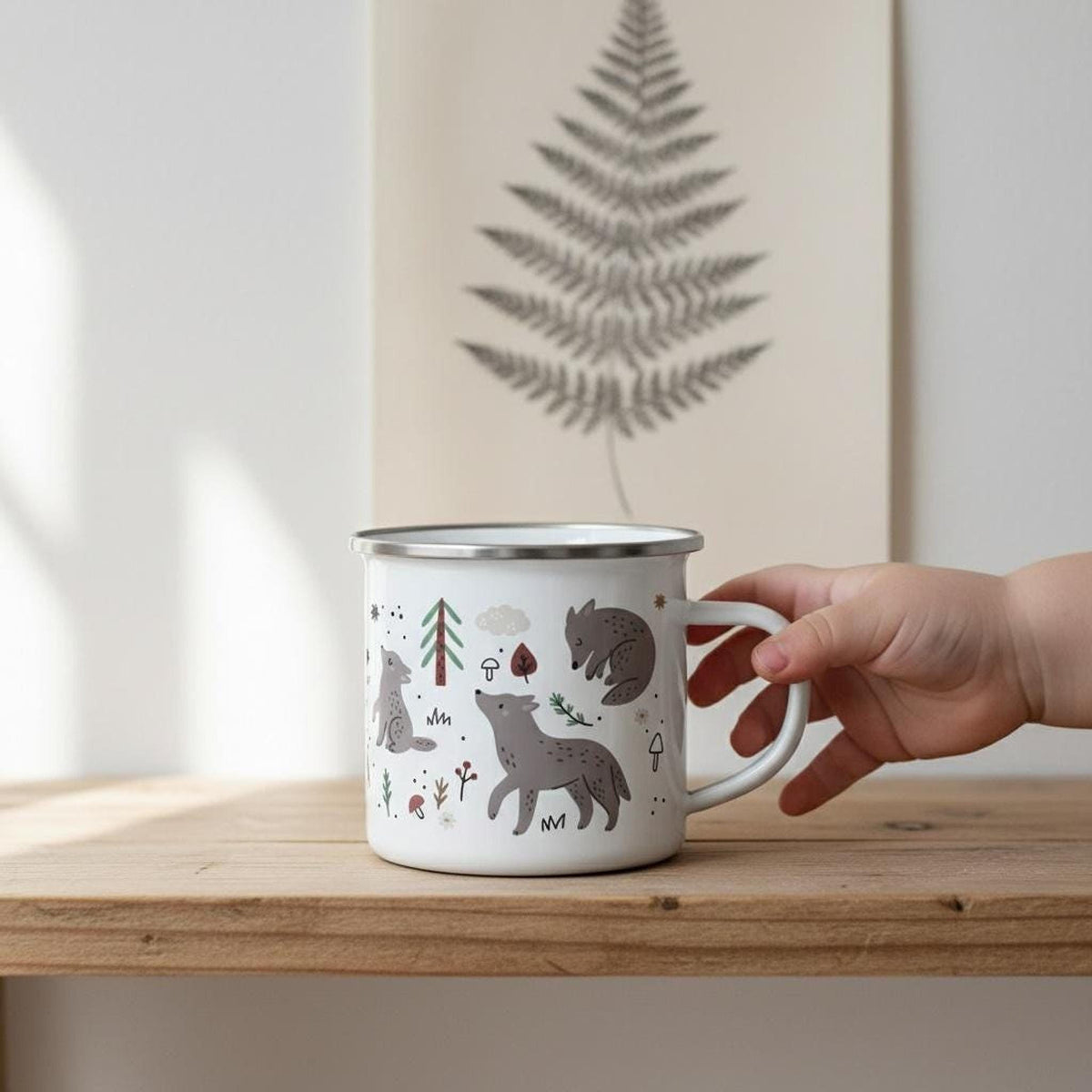 A child's hand holds a white enamel wolf mug on a wooden shelf next to a framed fern print.