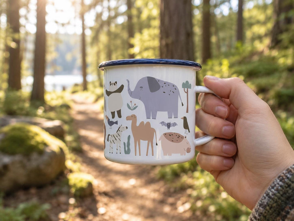 A white enamel mug with a dark blue rim featuring a colourful graphic print of various animals including an elephant, a camel, a panda, a zebra, a bat, and birds. The mug has a side handle and a smooth, glossy finish.