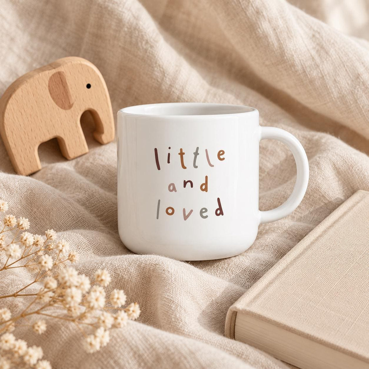 A white kids cup featuring the text "little and loved" printed in various muted earth-toned colours. The cup has a smooth, rounded handle and a cylindrical body. The text is arranged in three lines with a simple, sans-serif font.