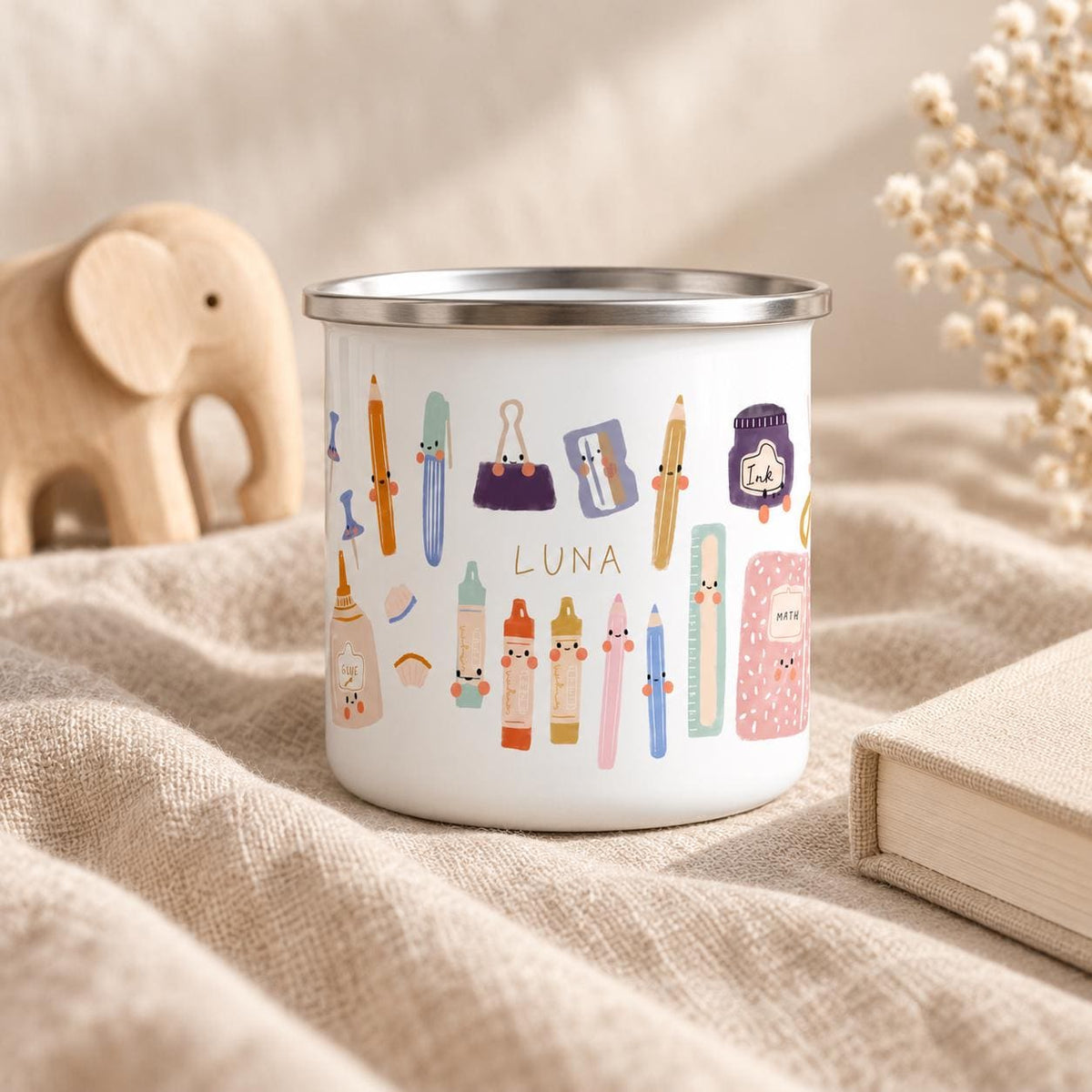 A white enamel mug with a silver rim featuring a colourful pattern of cartoon school supplies including pencils, crayons, a glue bottle, an ink pot, and a notebook. The name LUNA is printed in the centre among the illustrated items.