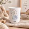 A white cup featuring a graphic of a grey rabbit standing on a blue moon with stars, a crescent moon, and a net. The name Noah is printed in black script at the bottom of the cup. The design includes soft blue, grey, and brown tones.