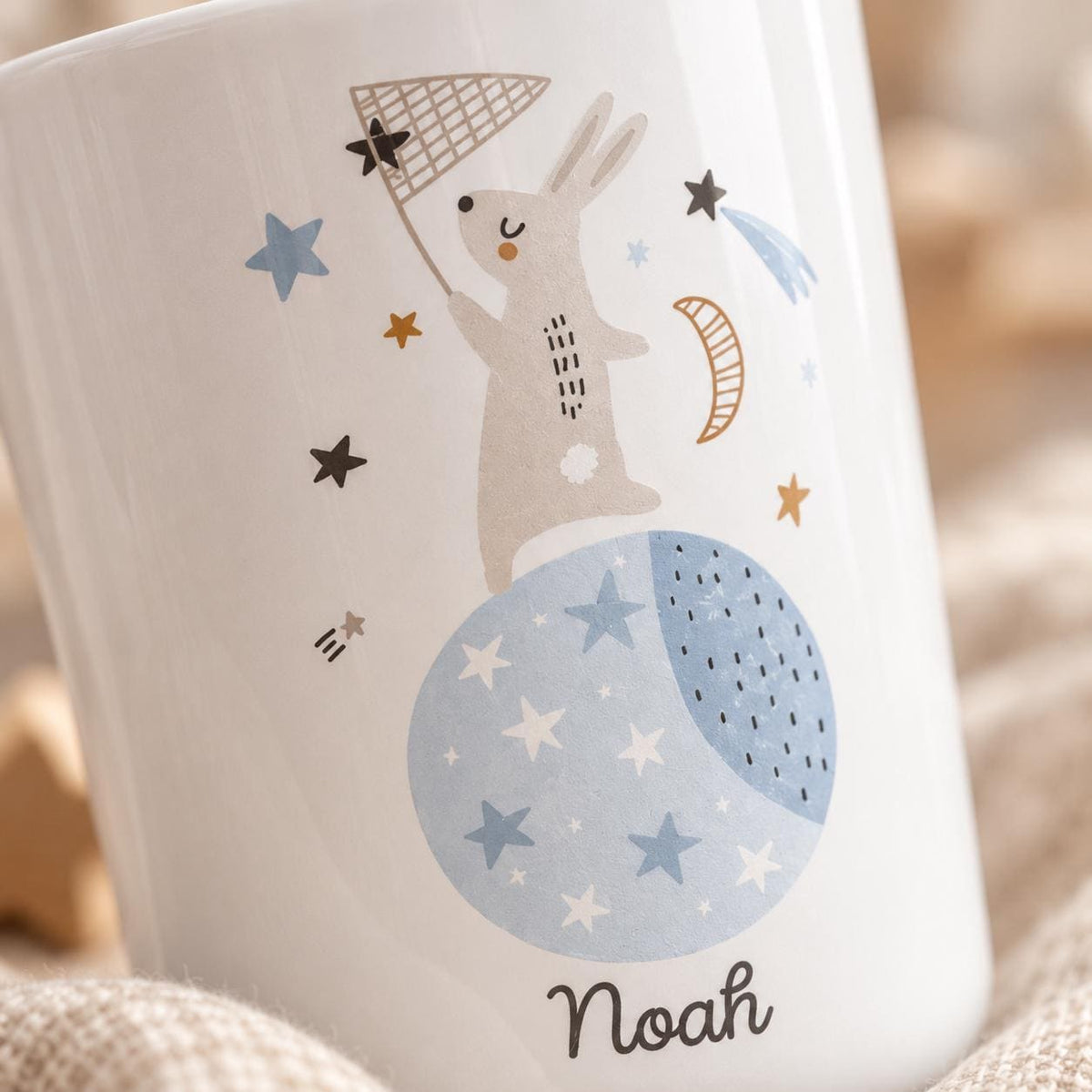A white kids cup featuring a graphic illustration of a light brown rabbit standing on a blue moon, holding a butterfly net. The design includes various stars, a crescent moon, and the name Noah printed in black script at the bottom.