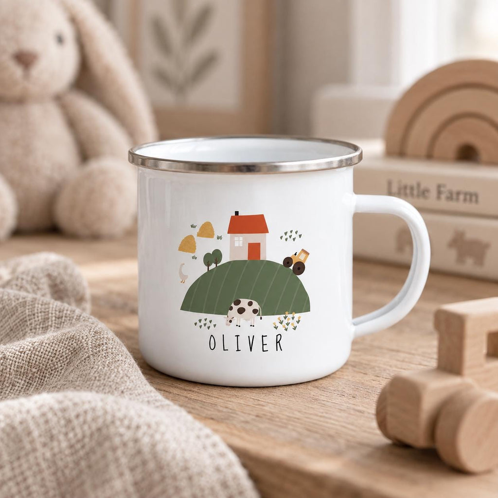 A white enamel mug with a silver rim and a handle, featuring a colourful farm scene illustration with a house, a cow, a tractor, and the name OLIVER printed in black capital letters on the front of the mug.