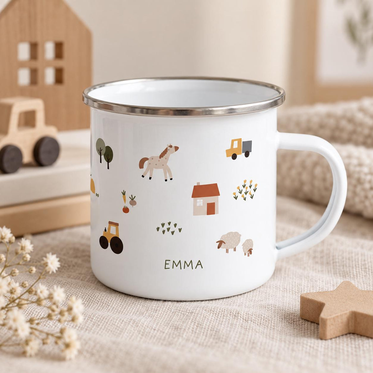 A white enamel mug with a silver rim and a handle, featuring a pattern of small illustrations including a horse, a tractor, a house, sheep, trees, and flowers. The name EMMA is printed in dark letters on the front of the mug.