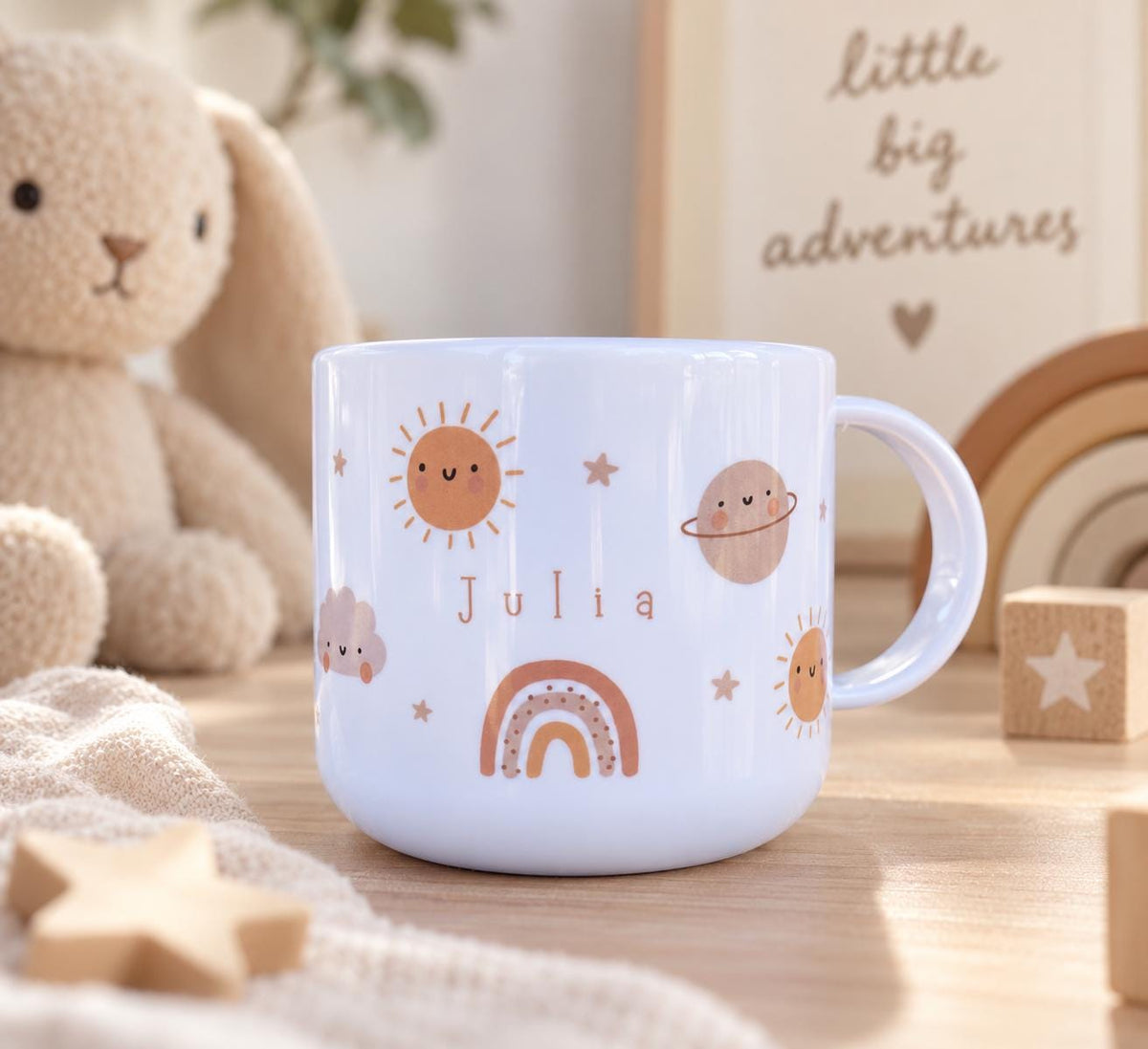 A white cup featuring a printed design of a smiling sun, a planet with rings, a small cloud, and a rainbow. The name Julia is printed in the centre in a brown sans-serif font. The cup has a smooth finish and a curved handle.