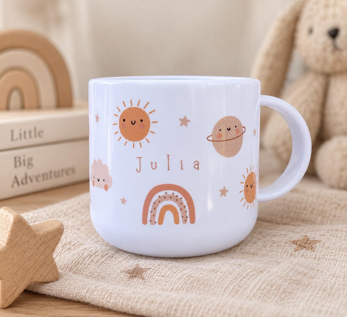 A white plastic cup featuring a printed design of a smiling sun, a planet with rings, a cloud, and a rainbow. The name Julia is printed in the centre in brown lettering. The cup has a single handle and a smooth, rounded exterior finish.