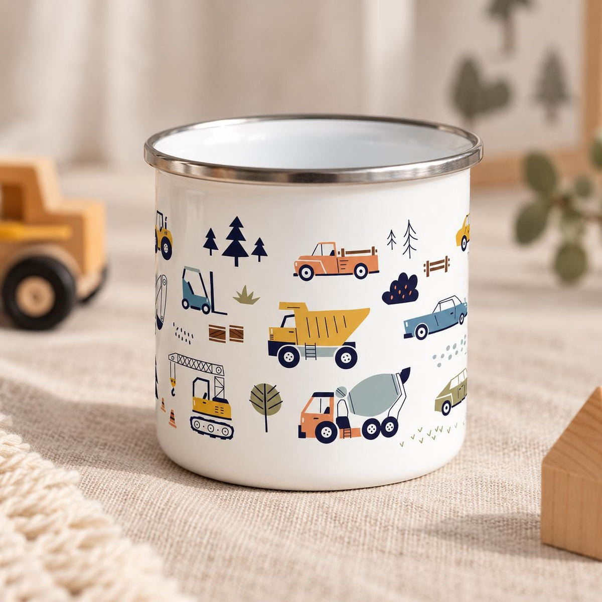 A white enamel mug with a silver-toned rim, featuring a repeating pattern of colourful construction vehicles, including dump trucks, cement mixers, and cranes, interspersed with small trees and bushes in shades of blue, yellow, and orange.