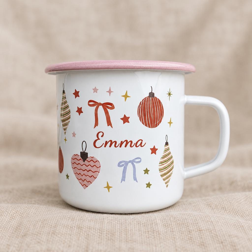 A white enamel mug with a pink rim and handle, featuring a festive pattern of red and gold Christmas ornaments, bows, and stars. The name Emma is printed in red script in the centre of the mug among the holiday decorations.