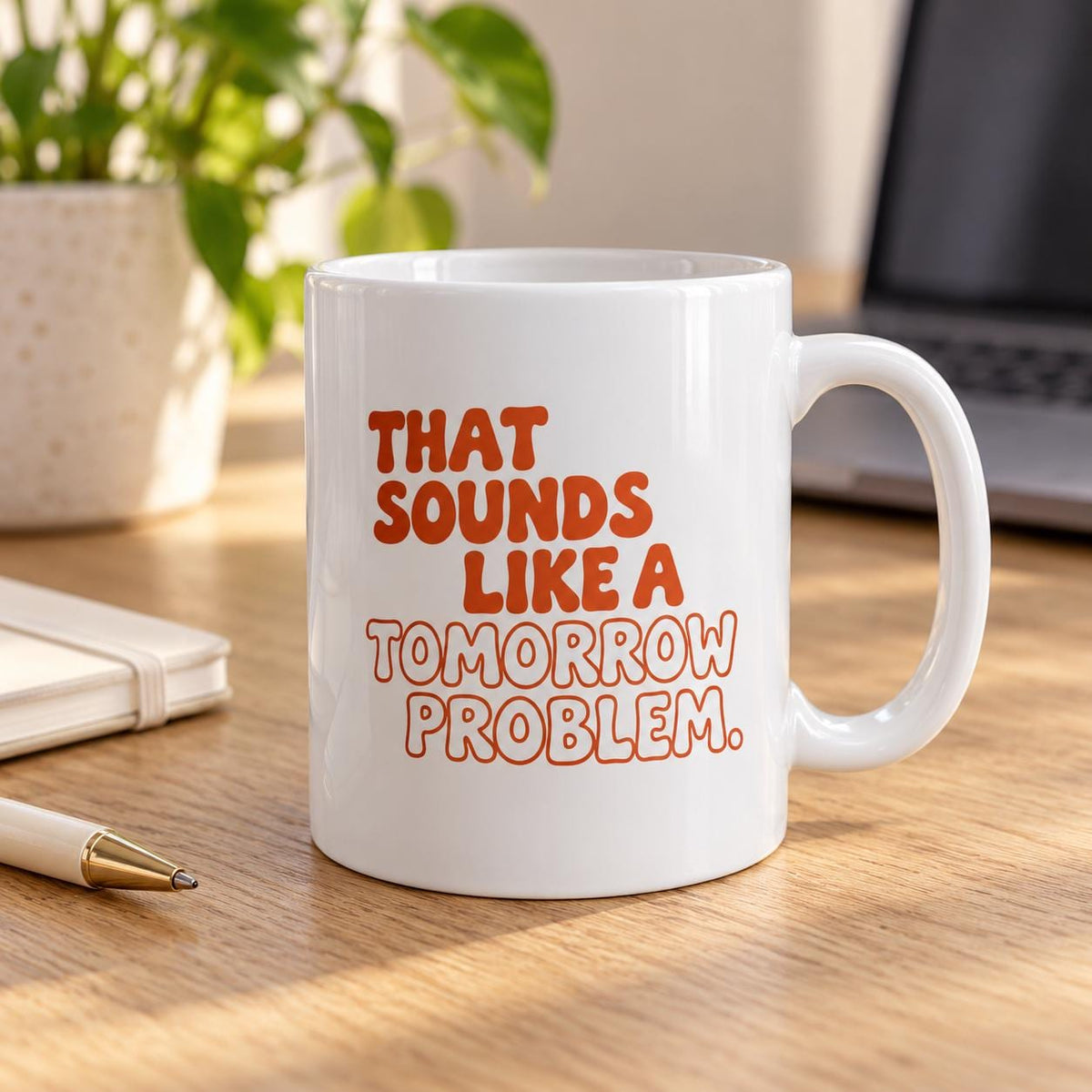 White ceramic That Sounds Like a Tomorrow Problem mug with bold burnt-orange lettering on a wooden desk beside a notebook, pen and plant.