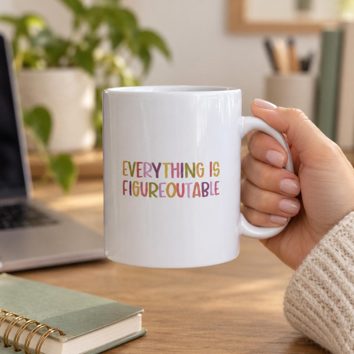 Hand holding the Everything Is Figureoutable mug at a desk with a laptop, notebook and plant.
