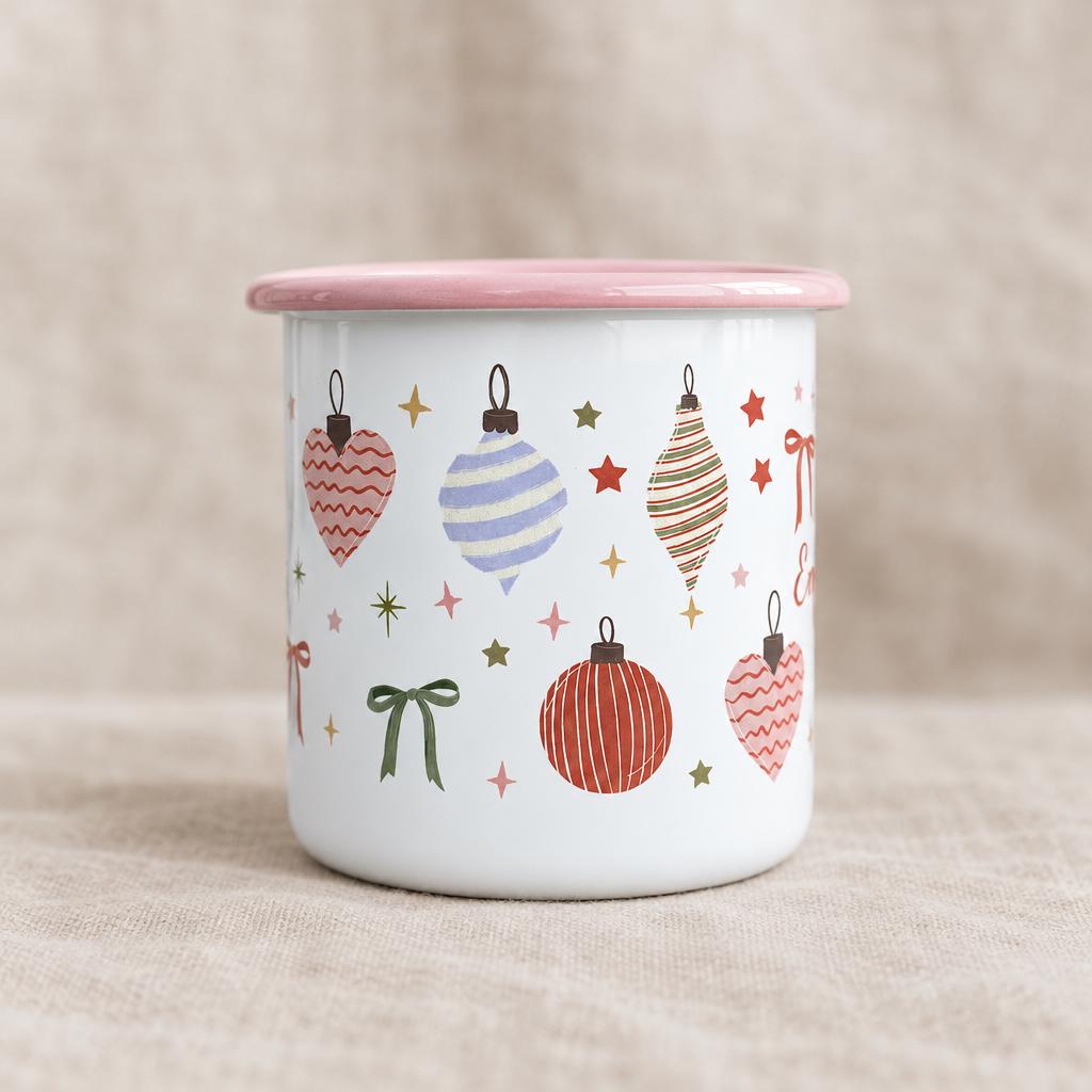 A white enamel mug with a pink rim featuring a repeating pattern of colourful Christmas ornaments, including heart-shaped baubles, striped spheres, green bows, and small stars in shades of red, green, pink, and blue.