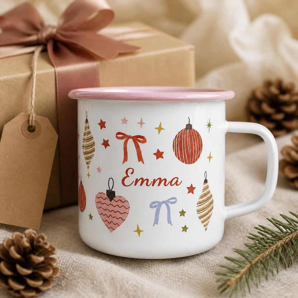 A white enamel mug with a light pink rim featuring a festive pattern of red and gold ornaments, bows, and stars. The name Emma is printed in red script in the centre. The mug has a side handle and a smooth, glossy finish.