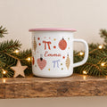 A white enamel mug with a pink rim and handle, featuring a festive pattern of red and gold Christmas ornaments, bows, and stars. The name Emma is printed in red script in the centre of the mug, surrounded by the colourful holiday graphics.