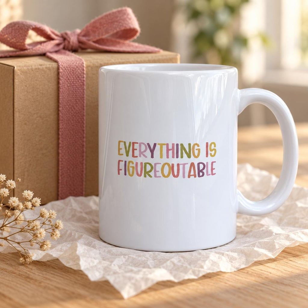 White ceramic Everything Is Figureoutable mug with colourful hand-lettered capitals beside a kraft gift box tied with a pink ribbon on a wooden table.