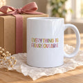 White ceramic Everything Is Figureoutable mug with colourful hand-lettered capitals beside a kraft gift box tied with a pink ribbon on a wooden table.