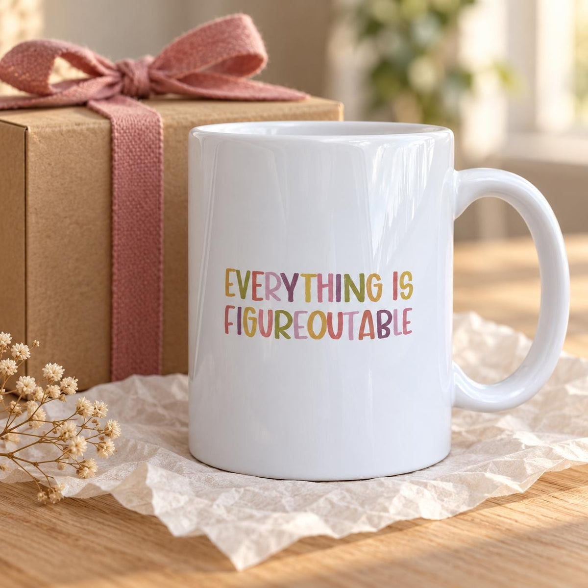 White ceramic Everything Is Figureoutable mug with colourful hand-lettered capitals beside a kraft gift box tied with a pink ribbon on a wooden table.