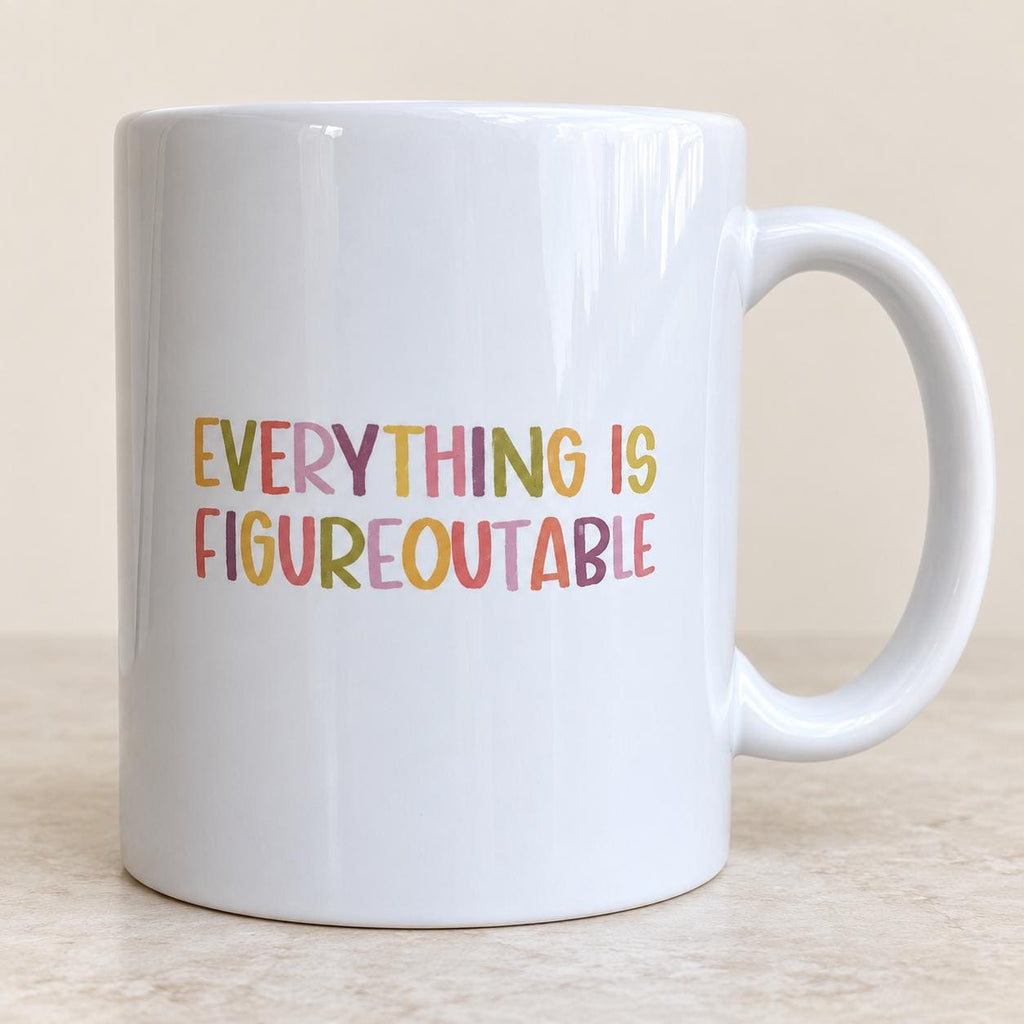 Everything Is Figureoutable mug with lettering in pink, mustard, purple and green, handle on the right, on a light stone surface.