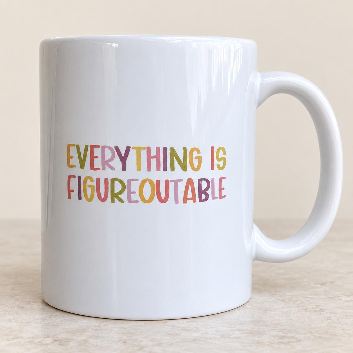 Everything Is Figureoutable mug with lettering in pink, mustard, purple and green, handle on the right, on a light stone surface.